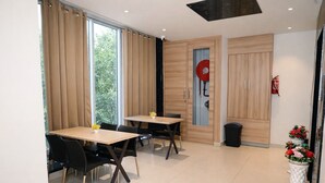 Un espace de restauration moderne avec deux tables et des chaises, de grandes fenêtres avec des rideaux, un extincteur et une poubelle.