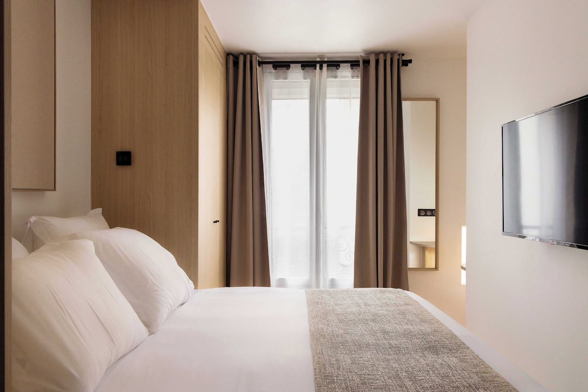 A hotel room with a large bed, a flat-screen TV mounted on the wall, and a window with curtains.