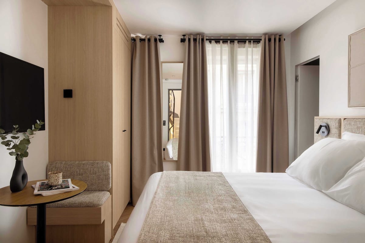 A hotel room with a bed, a small table with a vase and a magazine, a chair, and a TV mounted on the wall.