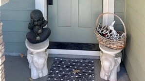 Une porte d’entrée surmontée d’une petite fenêtre, deux statues d’éléphants blancs de chaque côté, et un panier en osier avec une pancarte indiquant « Bonjour » sur la droite.