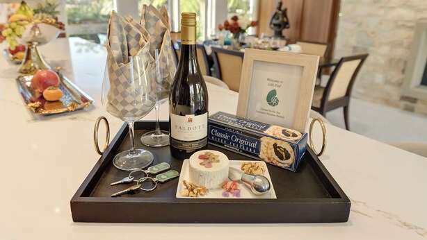 A tray with a bottle of wine, a cheese wheel, a box of crackers, a framed welcome sign, and a fruit platter. - 3954 Lilli Pad - Remodeled, Private Primary Suite (Carmel)
