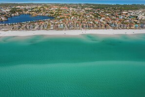 Luchtbeeld van een kustplaats met een strand, helder turquoise water en een mix van woon- en bedrijfsgebouwen.