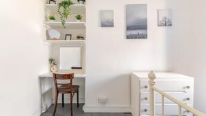 Ein Zimmer mit Schreibtisch, Stuhl und einem weißen Schminktisch. An der Wand befinden sich Regale mit Pflanzen und Kunstwerken.