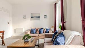 Ein Wohnzimmer mit einer Couch, einem Couchtisch und einer Pflanze. Vorhänge und eine Tür sind zu sehen.