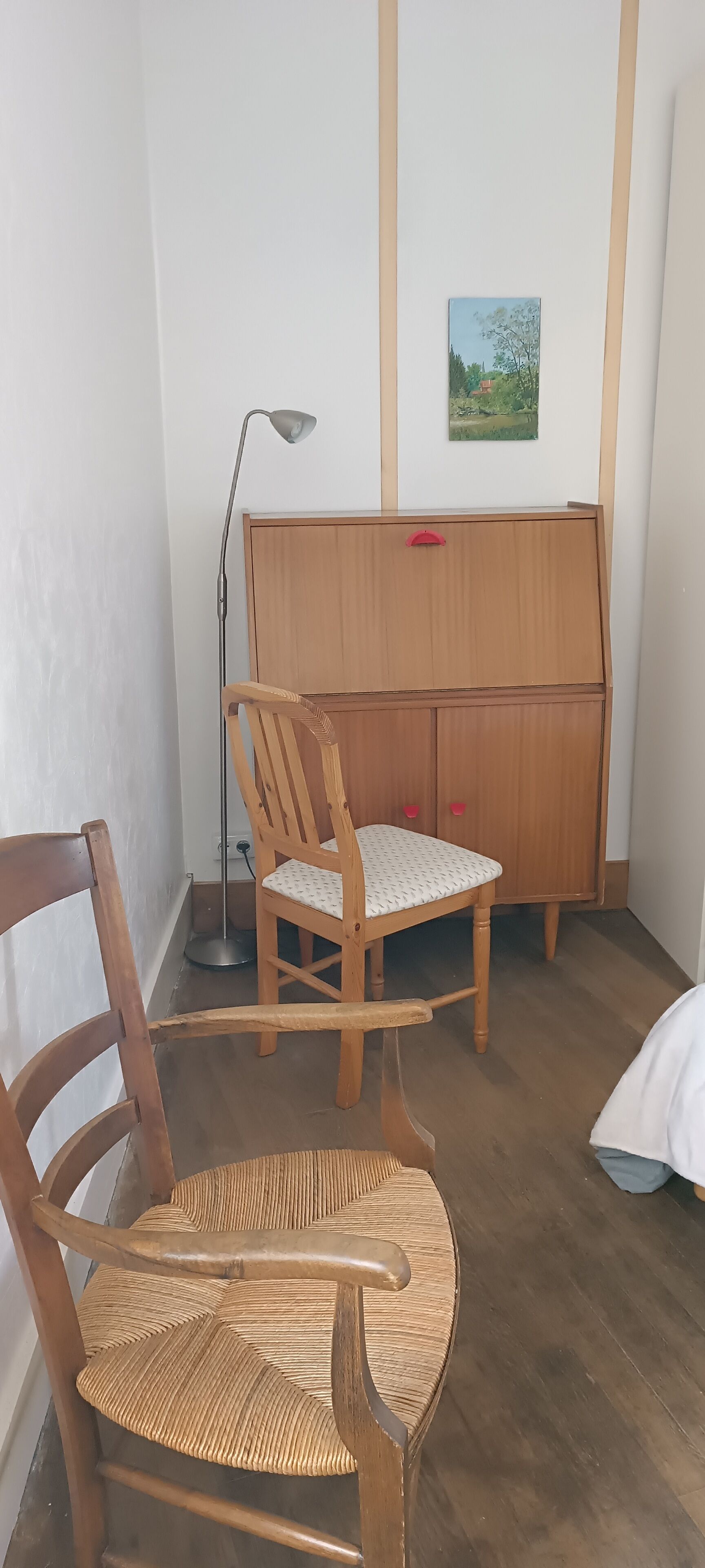 Une pièce d’angle avec un bureau en bois, une chaise, un lampadaire et un petit tableau accroché au mur.