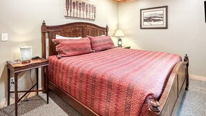 Une chambre avec un ventilateur de plafond en bois, un lit recouvert d’une courtepointe à motifs rouges, une table de chevet avec une lampe et un tableau encadré au mur.