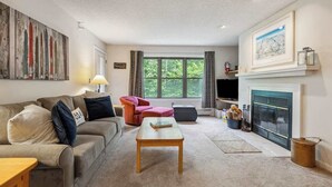 A cozy living room with a sofa, armchair, and coffee table, a fireplace, and a large window with curtains. - Mountain Escape 3 BR Condo at Okemo w/ Fireplace (Ludlow)