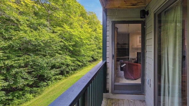 A wooden deck with a glass door leading to a room with a red chair and a kitchen visible inside. - Mountain Escape 3 BR Condo at Okemo w/ Fireplace (Ludlow)