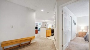 A hallway with a wooden bench, a kitchen area, and a bedroom with a nightstand and lamp. - Mountain Escape 3 BR Condo at Okemo w/ Fireplace (Ludlow)