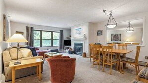 A living room with a sofa, armchair, coffee table, and dining table with chairs. There is a fireplace, a television, and a window with curtains. - Mountain Escape 3 BR Condo at Okemo w/ Fireplace (Ludlow)