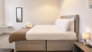 Ein ordentlich bezogenes Bett mit Kissen und einem gefalteten Handtuch, ein Nachttisch mit Lampe und ein kleiner weißer Tisch.