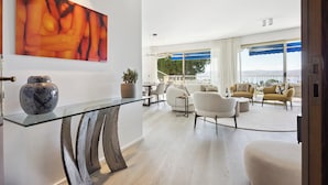 Un salon moderne avec une table basse en verre, un grand tableau abstrait et une vue sur l’extérieur grâce à de grandes fenêtres.