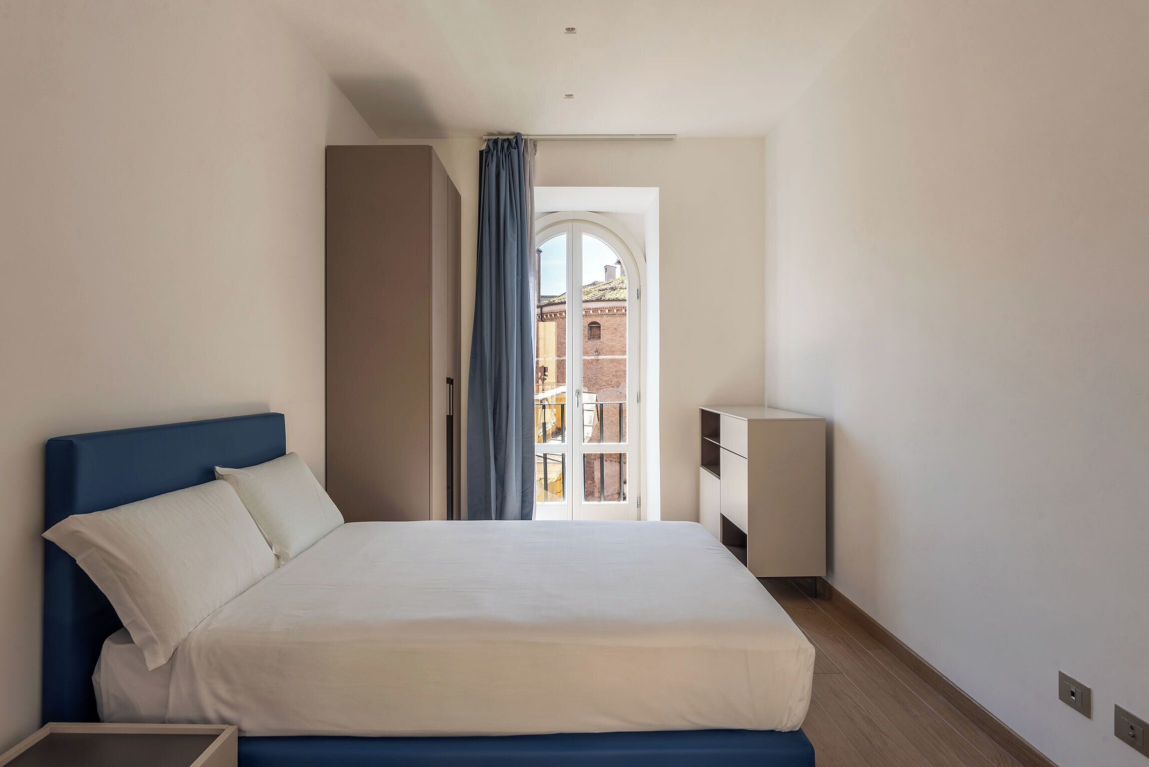 Une chambre à coucher avec un lit, une table de chevet, une commode et une vue sur un bâtiment par une porte ouverte.