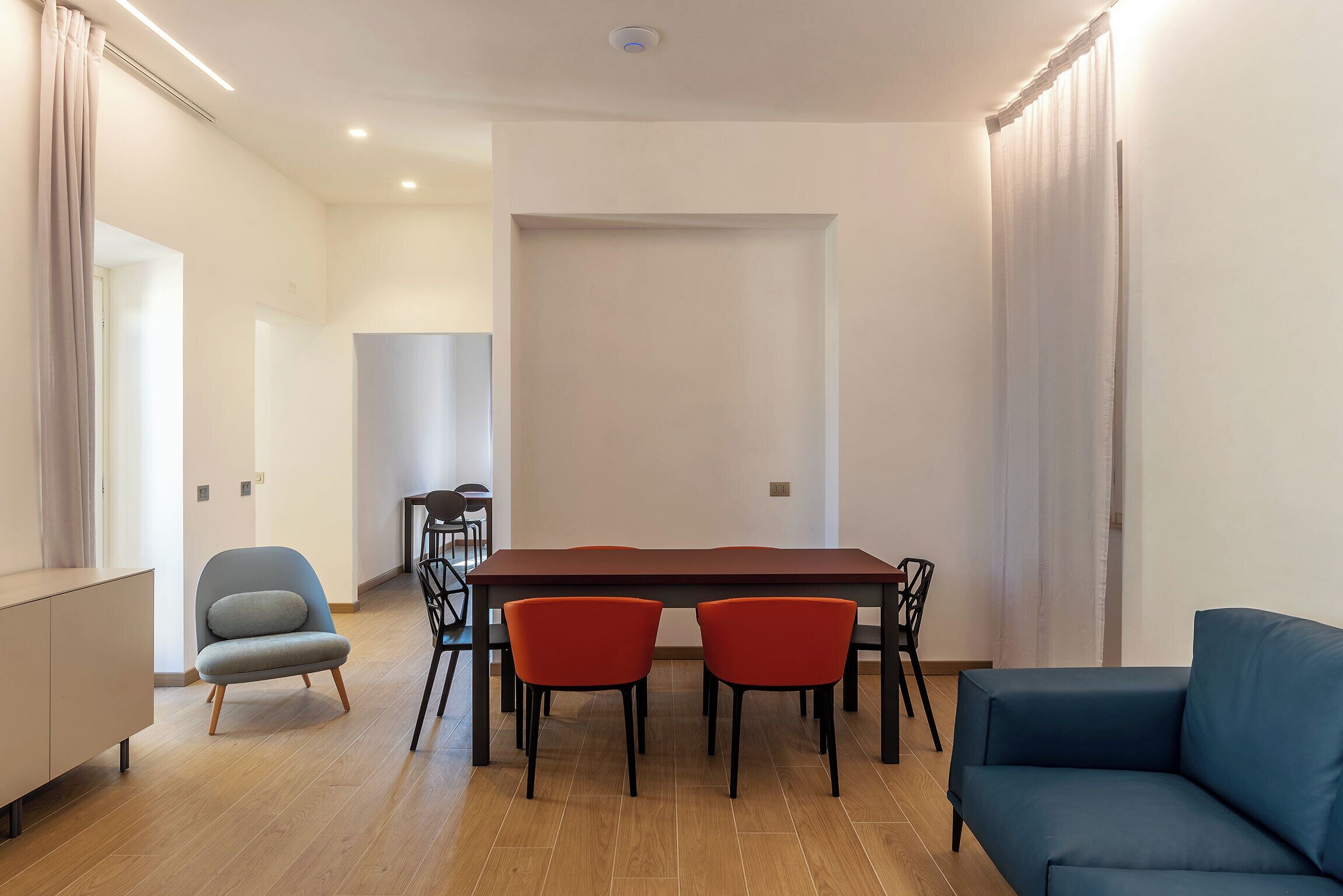 A modern living room with a dining table, two red chairs, a blue sofa, and a grey armchair.