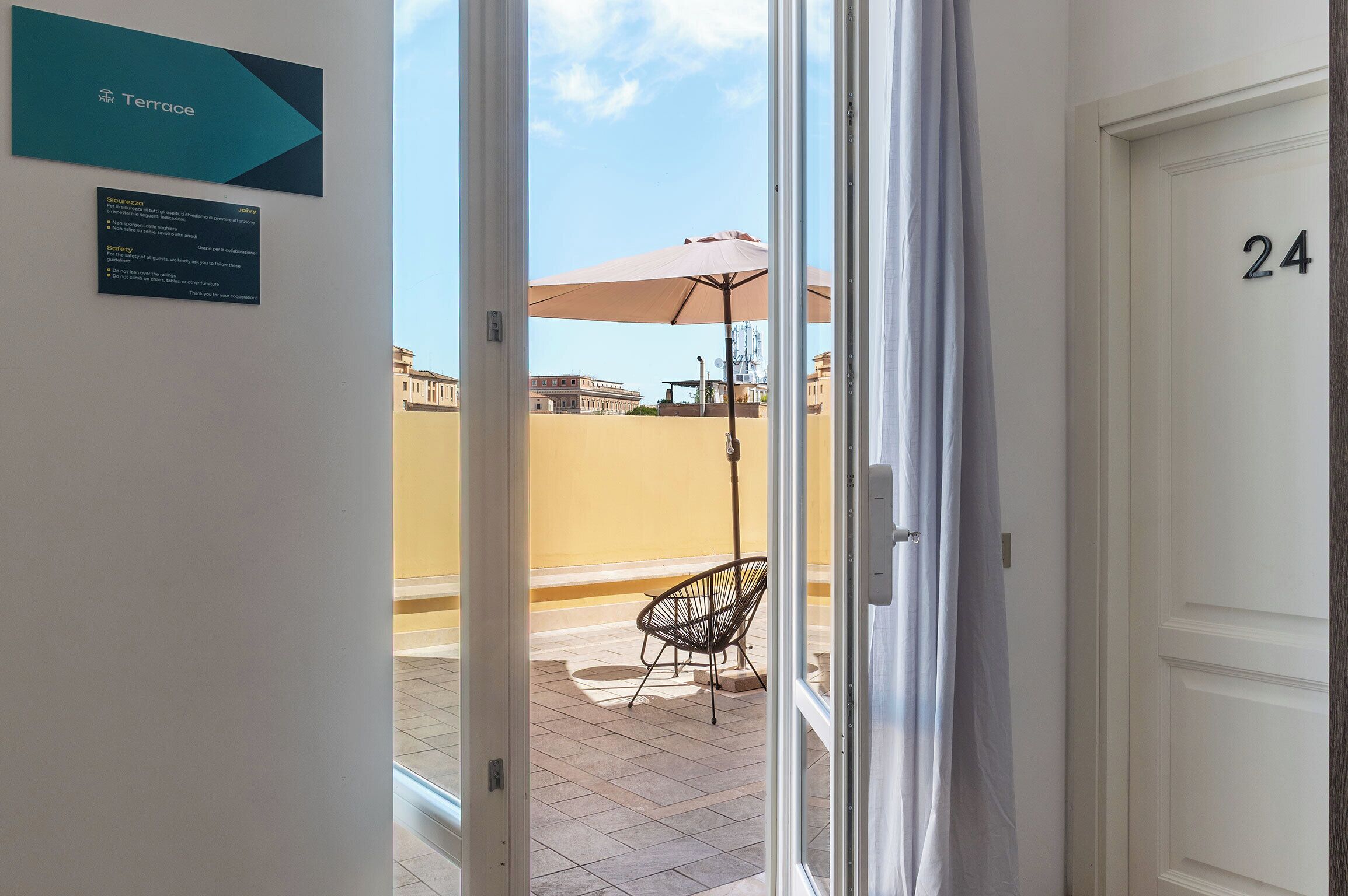 Entrada de hotel con un letrero que indica la habitación número 24, una terraza con una silla y sombrilla, y vistas a la ciudad.