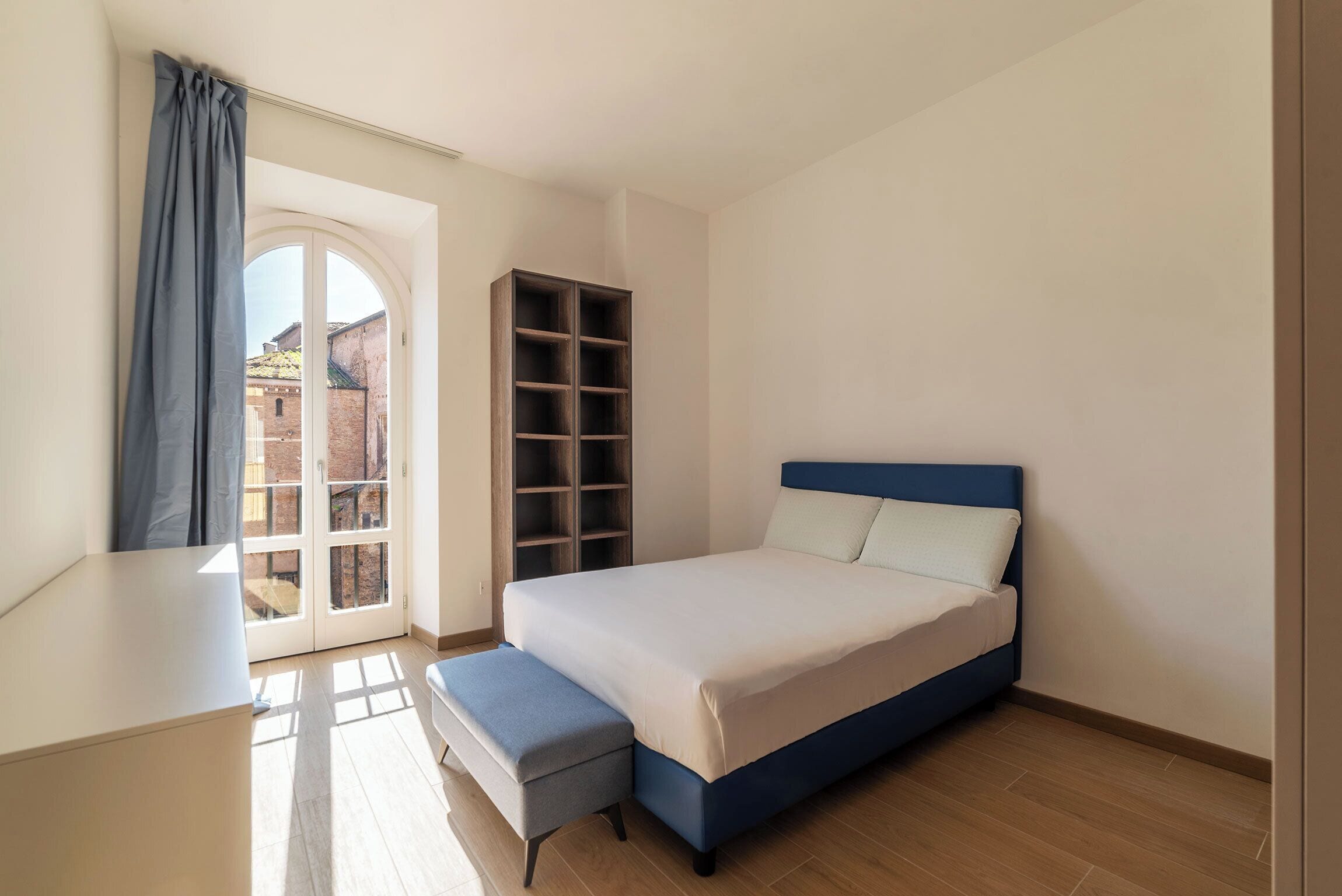 A bedroom with a bed, a bench, a wardrobe, and a view of a building through a glass door.