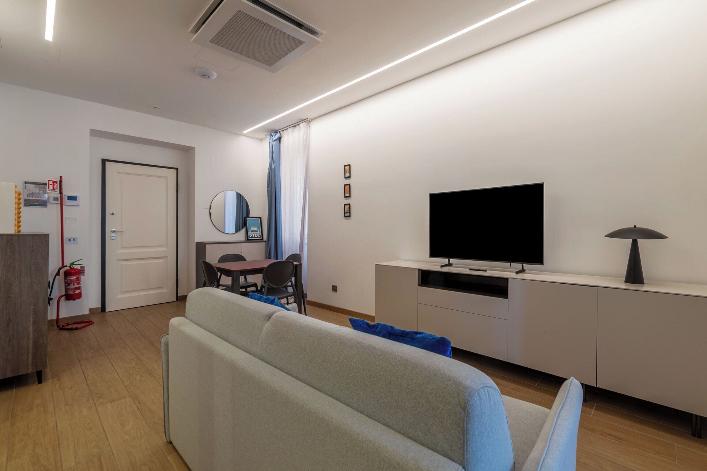 Ein modernes Wohnzimmer mit Sofa, Flachbildfernseher, Esstisch mit Stühlen, Feuerlöscher und einer Tür.