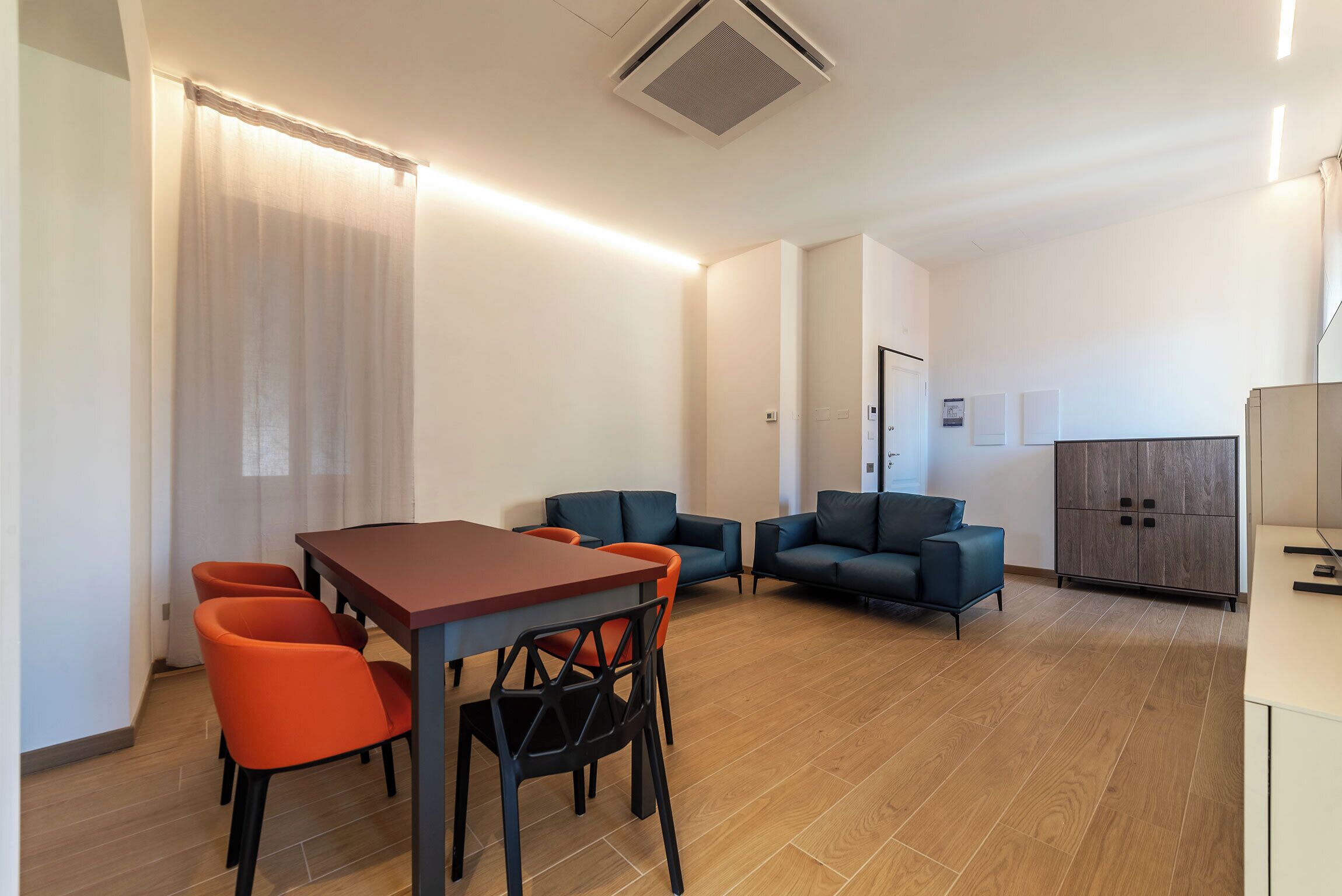 A modern, well-lit room with a wooden floor, a dining table with chairs, and two sofas.