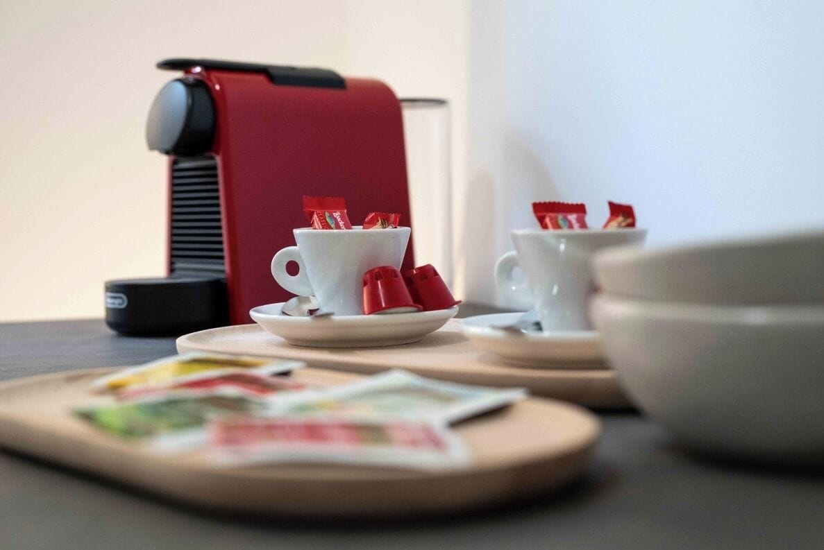 A red espresso machine, two white cups with red napkins, and a bowl on a table.