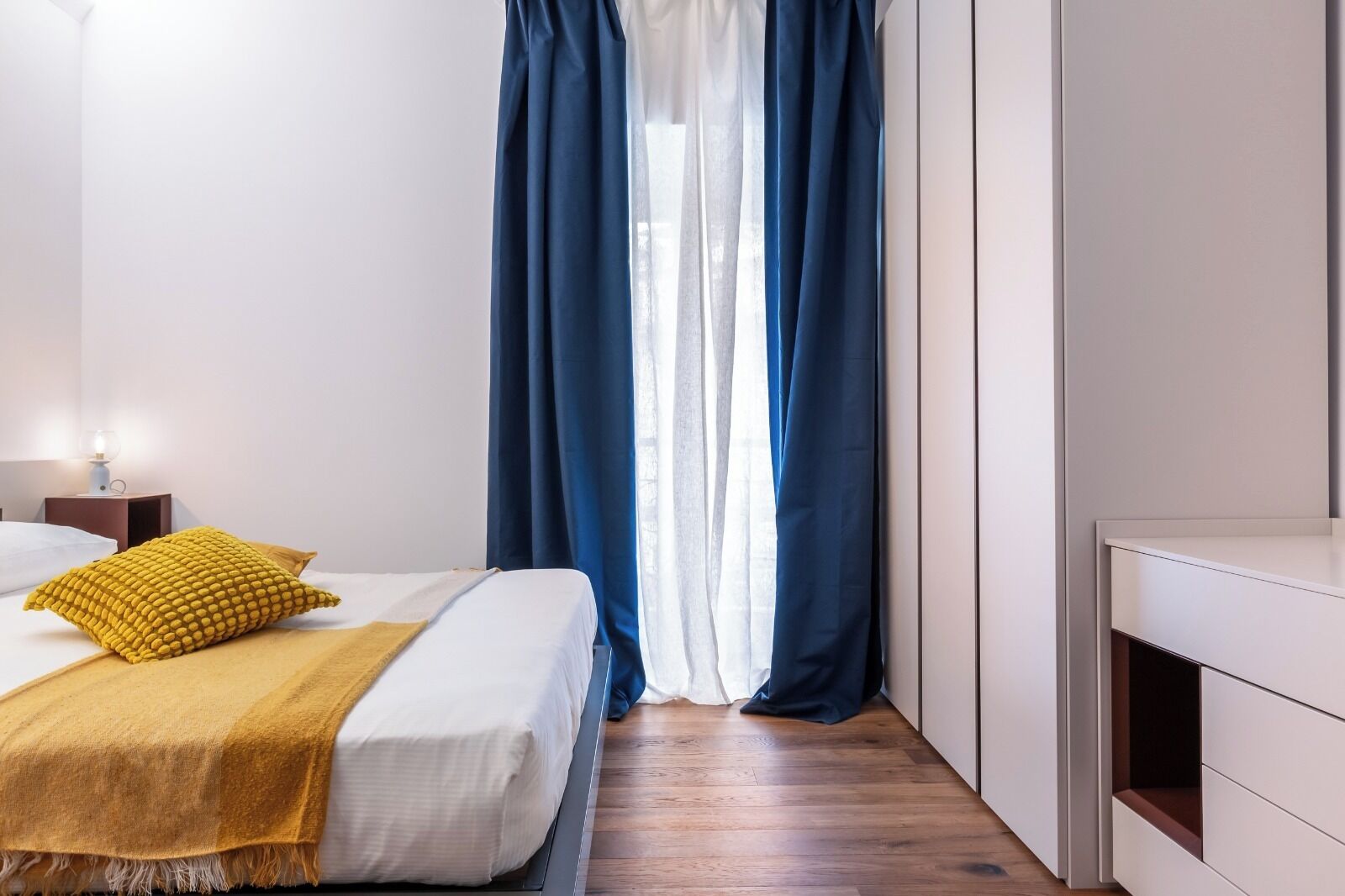Un dormitorio con cama, mesita de noche, lámpara y ventana con cortinas azules.