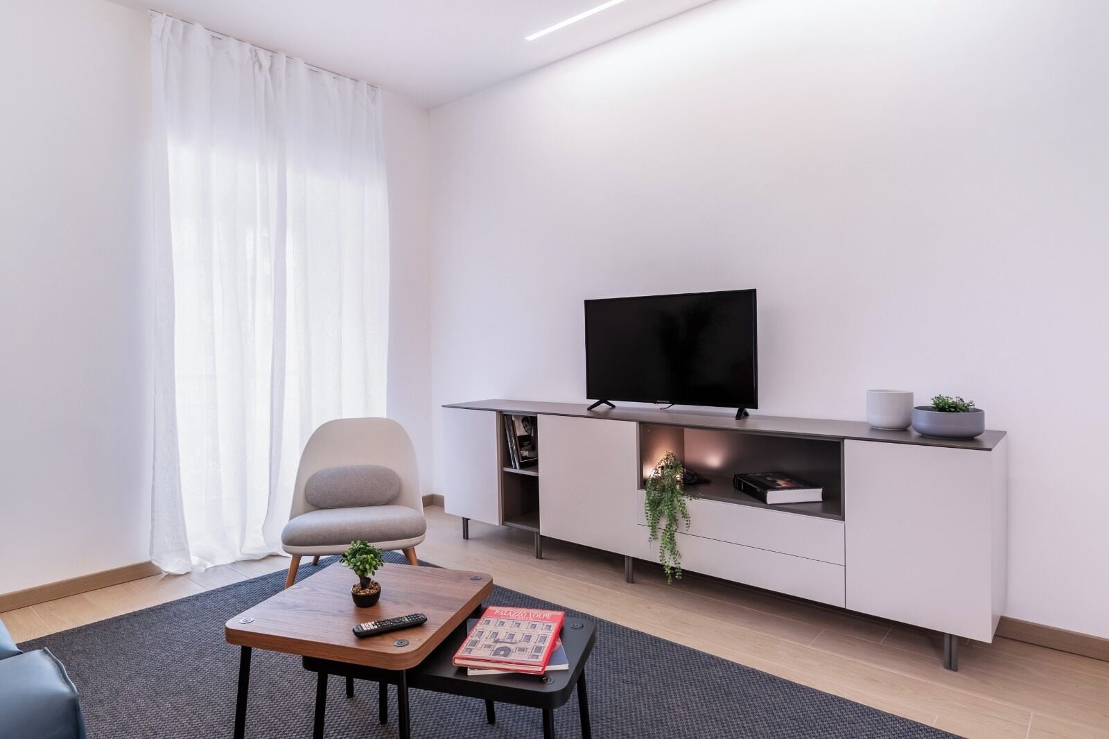 Un salon moderne avec un téléviseur à écran plat posé sur un meuble blanc, un fauteuil gris, une petite table basse avec une télécommande et un livre, et une plante en pot.