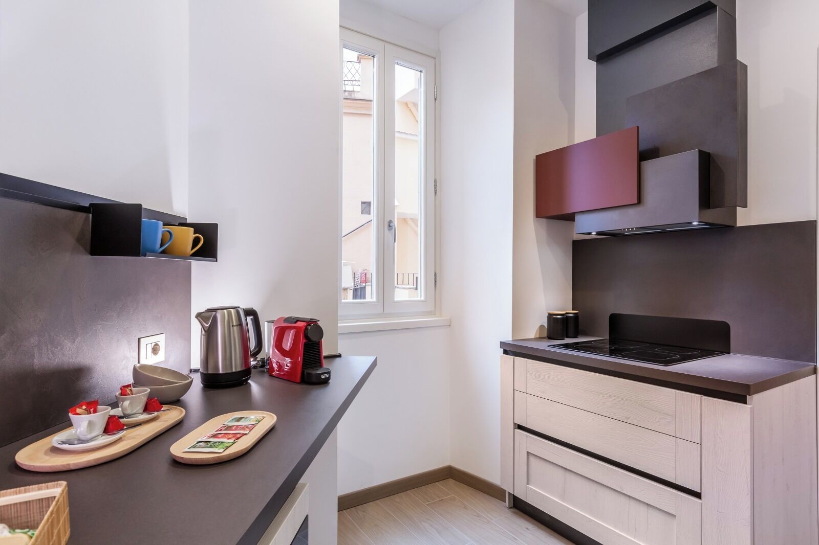 Una cocina moderna con encimera negra, hervidor de acero inoxidable, cafetera roja y tabla de cortar de madera con utensilios. Hay una ventana con vistas a edificios.