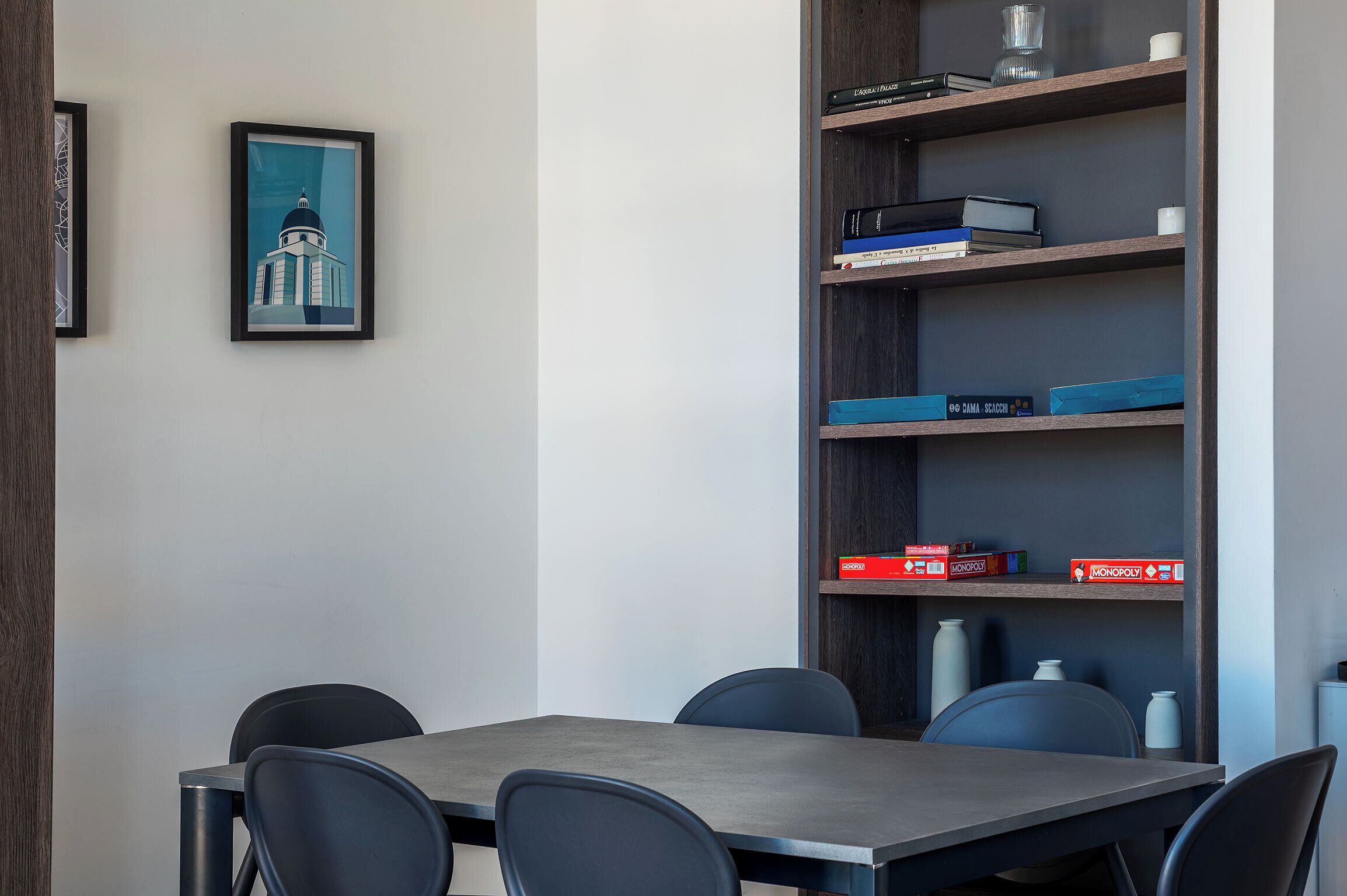 A modern room with a dining table, chairs, a bookshelf with books and board games, and a framed picture on the wall.