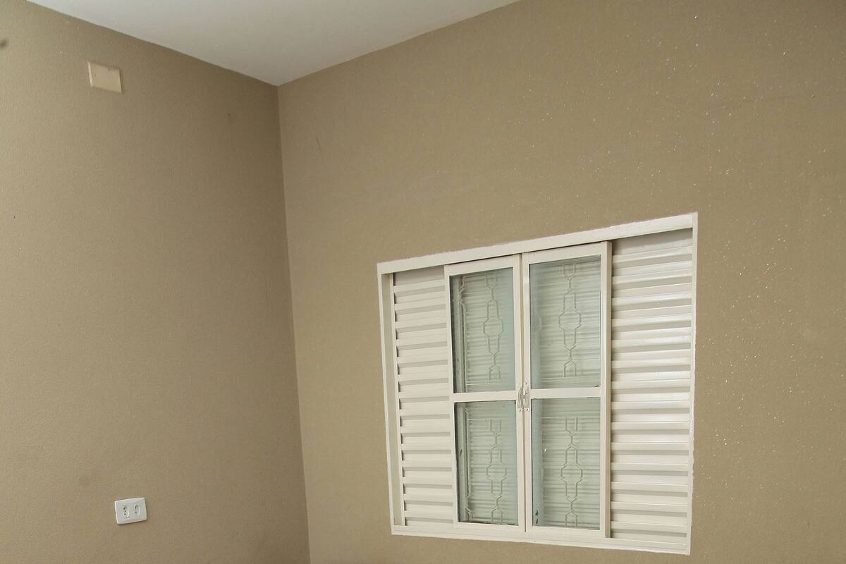 A room with two beds, each with a pink bedspread and white towels. There is a window with white shutters and a wall socket.