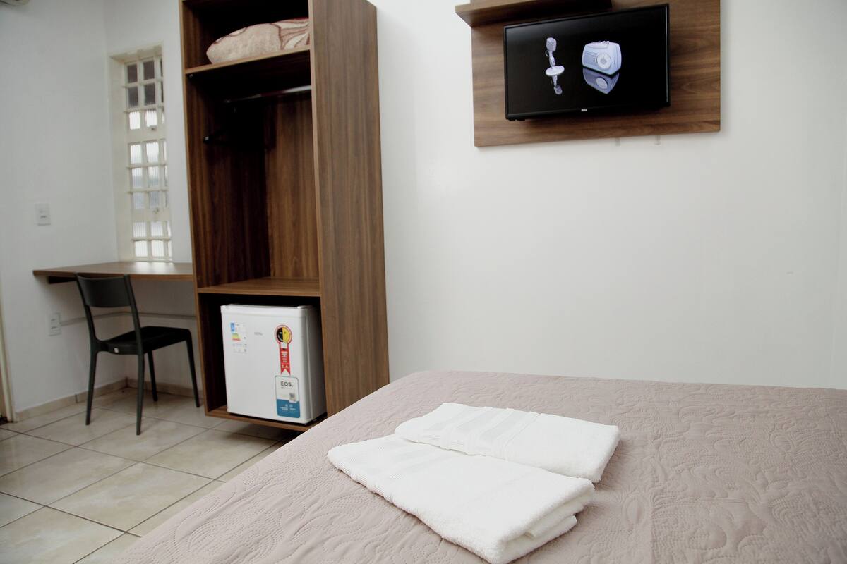 A bedroom with a bed, a wooden wardrobe, a desk with a chair, and a television mounted on the wall.