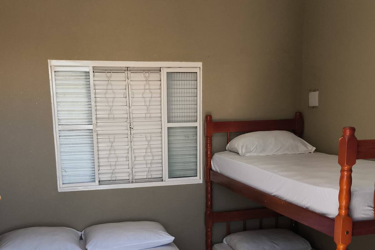 A dormitory room with bunk beds, a window with blinds, a bed with a white sheet and pillows, and a tiled floor.