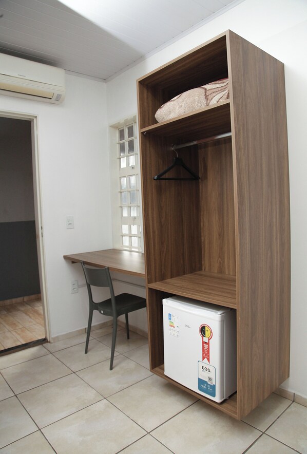 A compact room with a wooden desk, a chair, a white refrigerator, and a window with blinds. - HOTEL MODELO (Ribas do Rio Pardo)