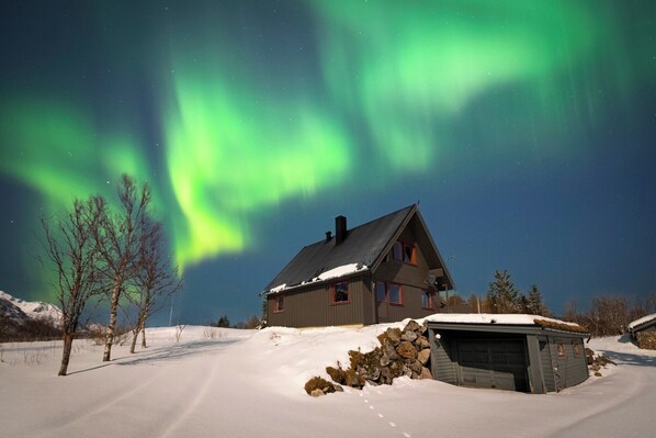 A house with a snow-covered roof and a garage, surrounded by a snowy landscape under a night sky with visible stars. - Bestefarhaugen - the Cozy House on the Hill,& Dome (Bø)