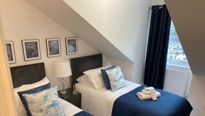Een hotelkamer met twee bedden, elk voorzien van een blauwe sprei en bijpassende kussens. Boven de bedden hangen prestatiekaders, er is een lampje op het nachtkastje en een raam met gordijnen.