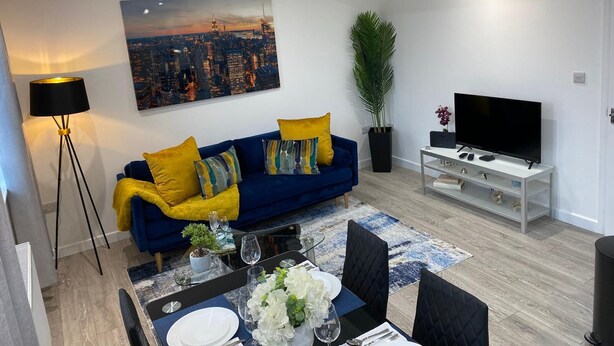Een moderne woonkamer met een blauwe bank, gele kussens, een stadsgezicht schilderij, een potplant, een televisie op een witte kast, en een eethoek met een tafel gedekt voor twee personen.