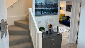 Een trap met beklede treden en een witte leuning, die leidt naar een kamer met een schilderij van een berglandschap, een aan de muur bevestigd telefoon en een klein bureau met een agenda en decoratieve objecten.