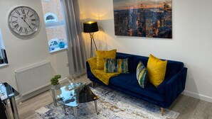 Een moderne woonkamer met een blauwe bank, gele kussens, een stadslandschap schilderij, een ronde wandklok en een vloerlamp.