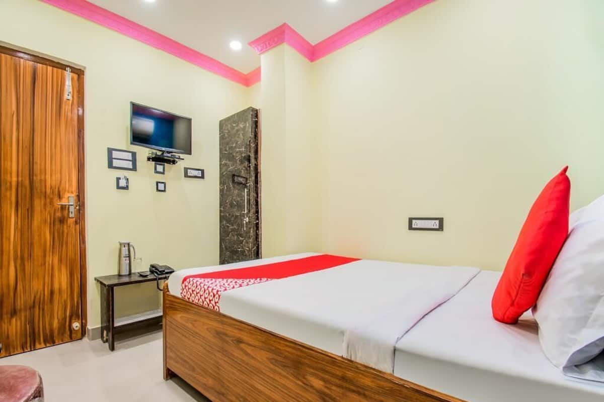 A hotel room with a wooden bed, a television, a small table with a kettle, and a red pillow.