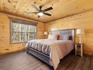 A wooden cabin bedroom with a bed, two bedside lamps, a ceiling fan, and a window with a view of trees. - NEW Luxury GBurg Cabin Private Pool-Theater-Games- Hot Tub (Gatlinburg)