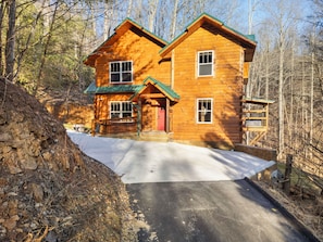 A two-story log cabin with a green roof, a front porch, and a snow-covered driveway.