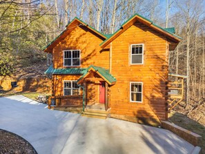 A two-story log cabin with a green roof, a front porch, and a visible garage door. - NEW Luxury GBurg Cabin Private Pool-Theater-Games- Hot Tub (Gatlinburg)