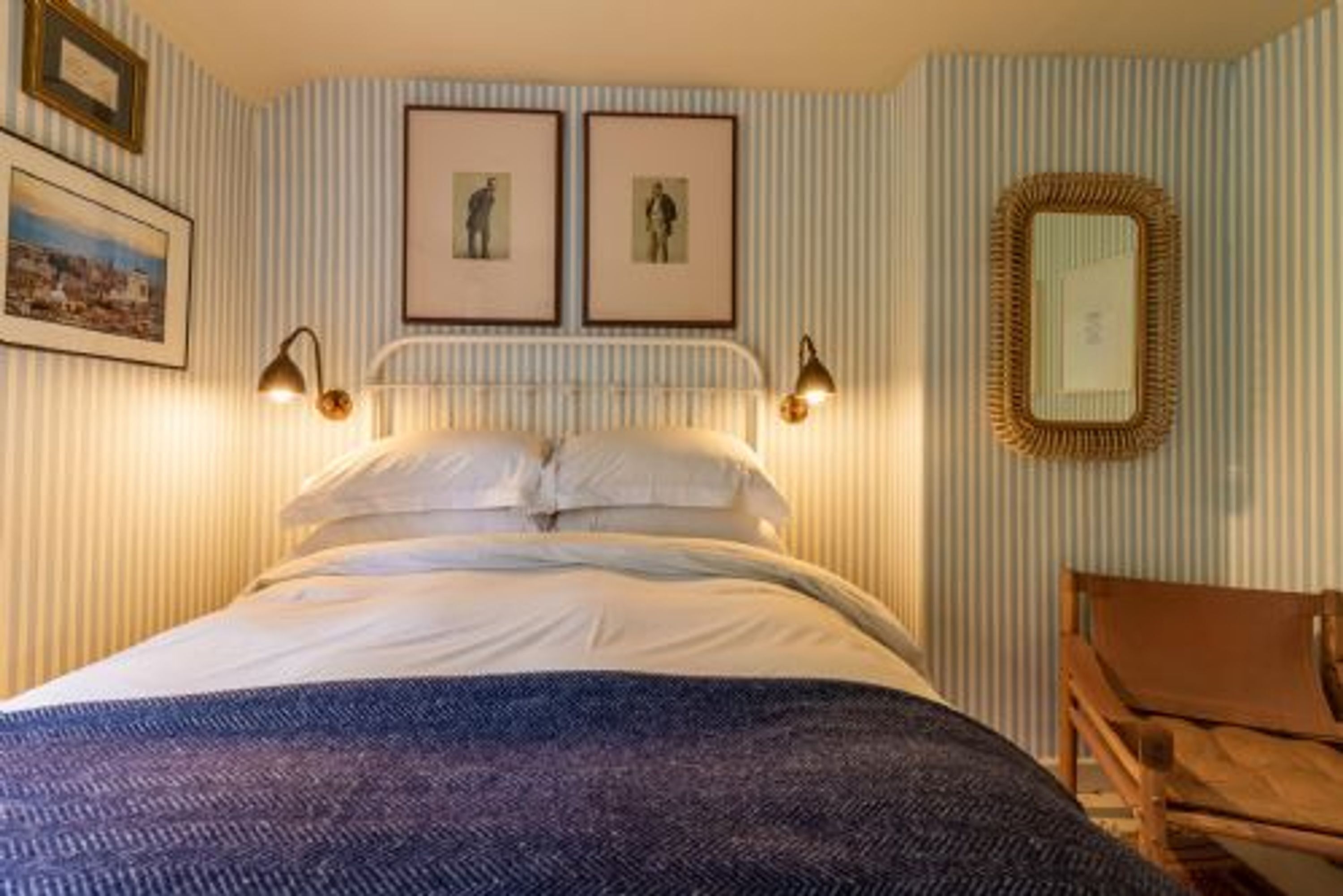 A neatly made bed with a patterned blanket, two framed pictures on the wall, and a chair to the right.