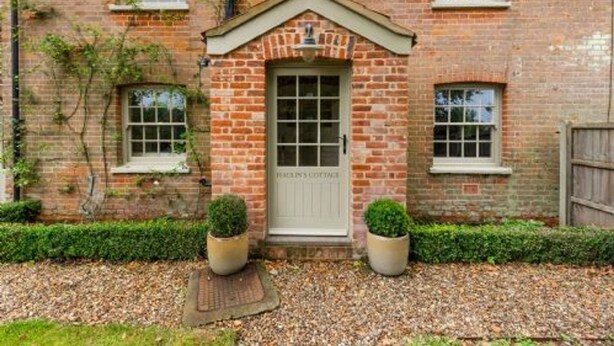A brick cottage with a white door labeled "HALLIN'S COTTAGE," two potted plants, and a gravel driveway. - Host Stay Haclins Cottage (Fakenham)