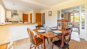A dining area with a wooden table set for four, surrounded by wooden chairs. A kitchen with white cabinets and a wooden door is visible in the background. - Host Stay Welsford House (Norwich)