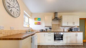 A kitchen with white cabinets, a large clock, and a tiled backsplash. - Host Stay The Hideaway (Wells-next-the-Sea)