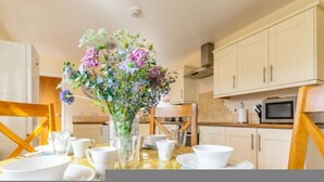 A dining area with a table set for two, a vase of flowers, and a kitchen in the background. - Host Stay The Hideaway (Wells-next-the-Sea)