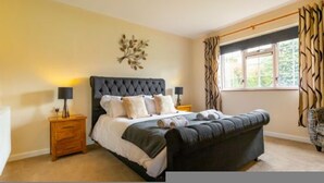 A bedroom with a large bed, two bedside tables, a wall-mounted light fixture, and a window with curtains. - Host Stay The Hideaway (Wells-next-the-Sea)
