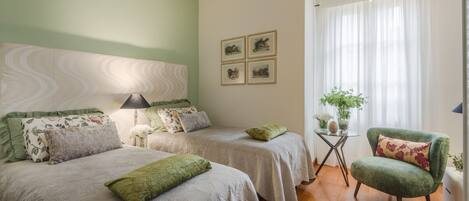 A bedroom with two beds, a green armchair, a small table with a plant, and a wall-mounted air conditioner.