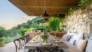 Un espacio de comedor al aire libre con una larga mesa de madera dispuesta para una comida, rodeada de sillas y un banco acolchado.