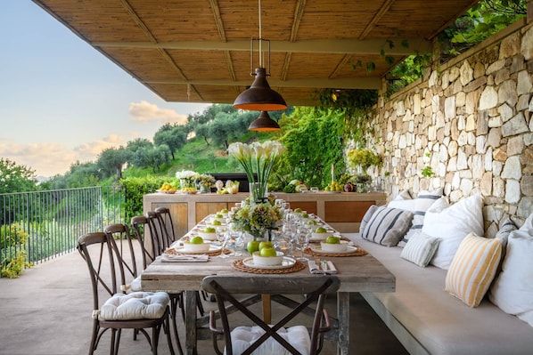 Un espace de restauration en plein air avec une longue table en bois dressée pour un repas, entourée de chaises et d’un banc rembourré.