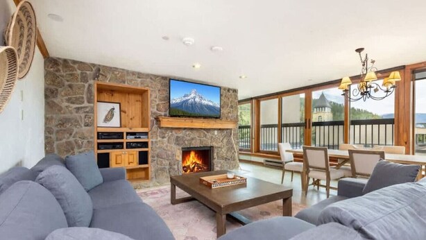 A living room with a stone fireplace, a television, and a balcony with outdoor seating.